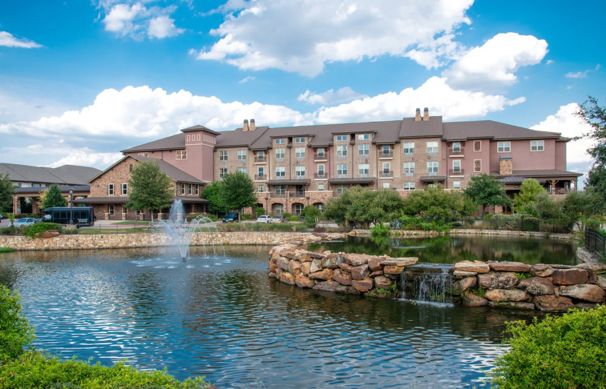 A scenic view of a hotel by a lake with fountains and lush greenery under a partly cloudy sky.