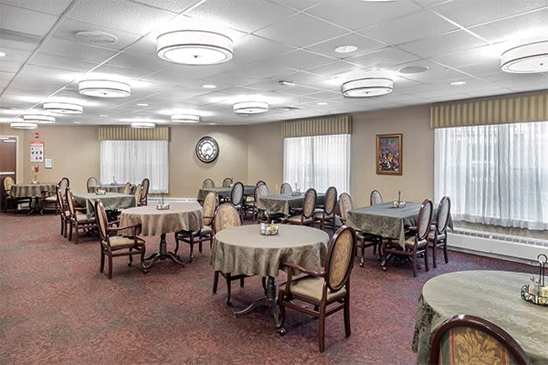 A well-lit dining area with multiple round tables and upholstered chairs arranged on a carpeted floor.