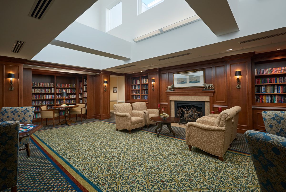 A cozy library room featuring armchairs, a fireplace, and bookshelves filled with books.