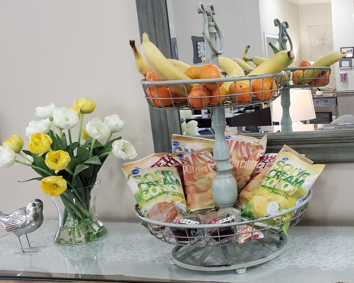 A decorative table display featuring a two-tiered stand with bananas, oranges, and various flavored chips, alongside a vase of fresh yellow and white flowers.