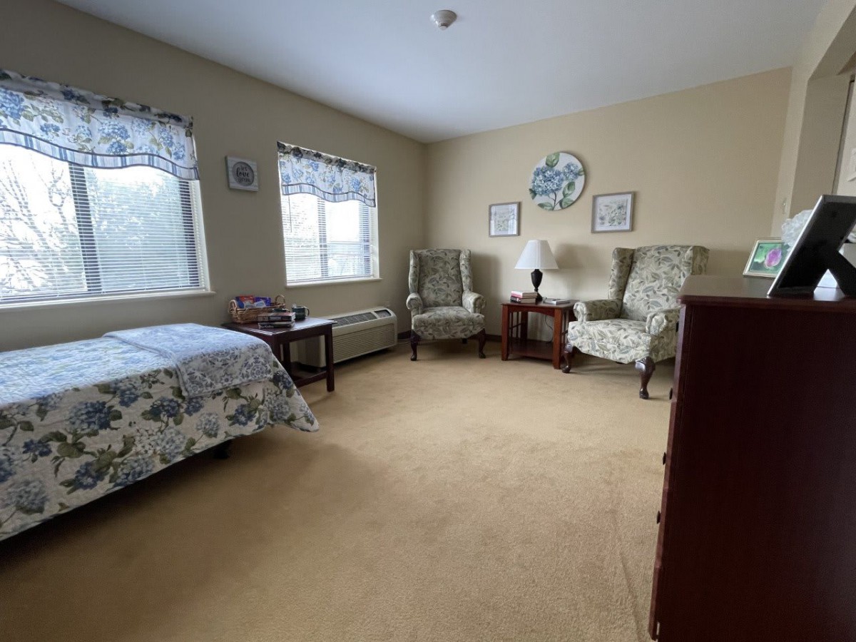 A cozy bedroom with floral decor, featuring two armchairs, a bed, and windows allowing natural light.