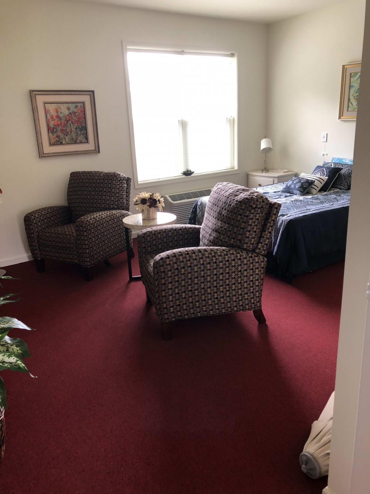 A cozy room with two upholstered chairs, a small table, and a bed near a large window.