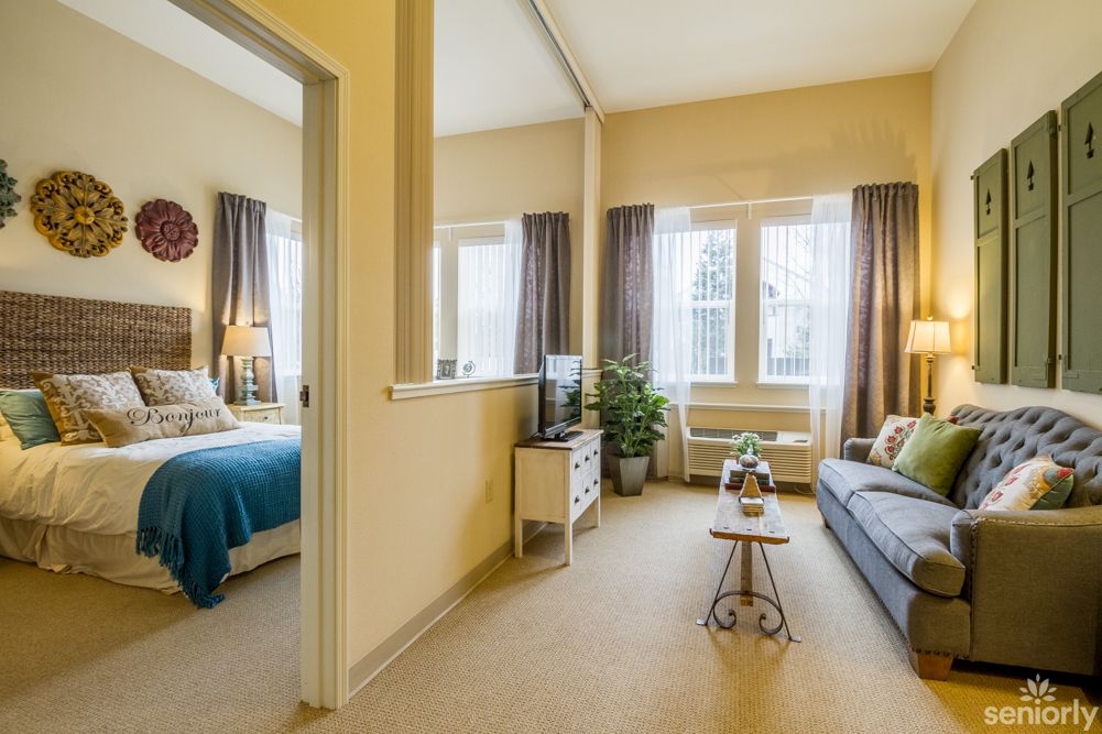 A cozy apartment interior featuring a living room with a sofa and a bedroom visible through an open doorway, illuminated by natural light from large windows.