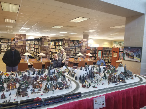 A festive miniature train and village display set up in a library, surrounded by bookshelves.