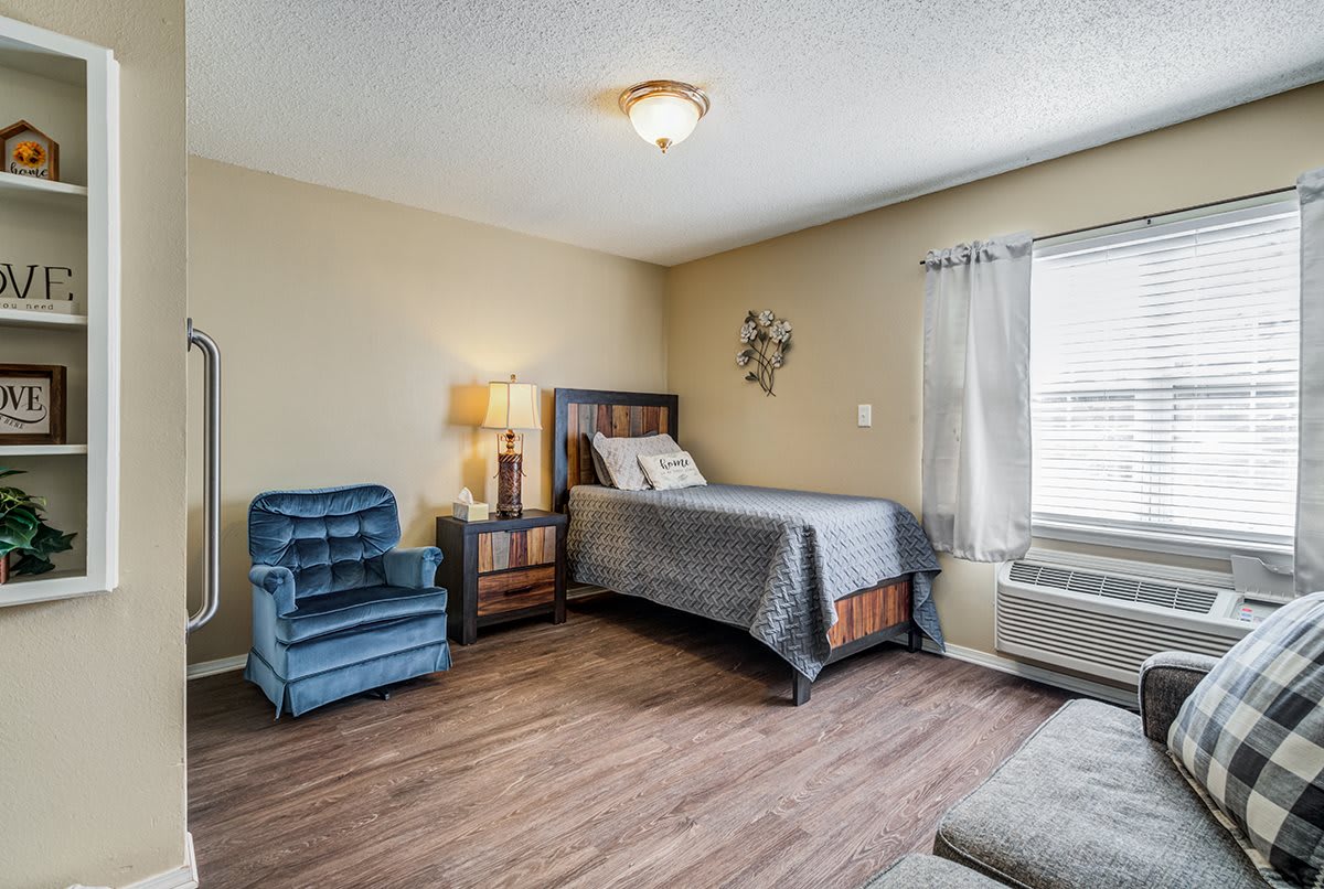 A cozy bedroom with a single bed, blue chair, nightstand, and window with curtains.
