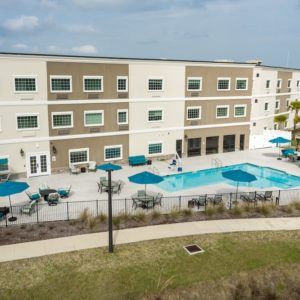 An apartment complex with a swimming pool area surrounded by tables and umbrellas, enclosed by a fence.