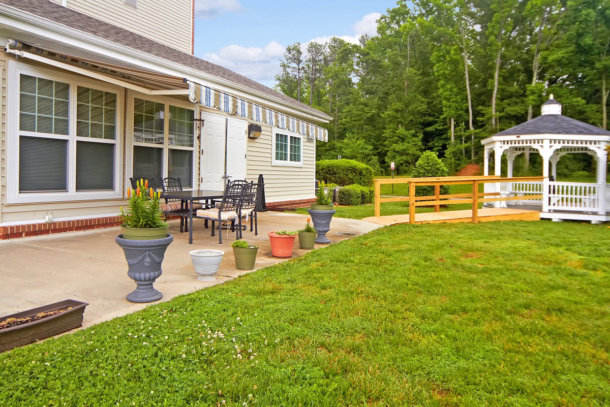 A backyard patio area with outdoor furniture, surrounded by a lawn and garden, featuring potted plants and a gazebo in the background.
