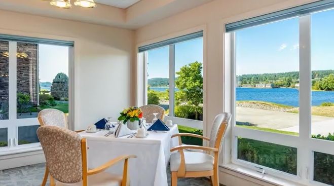 A cozy dining area with a beautifully set table, overlooking a scenic waterfront through large windows.