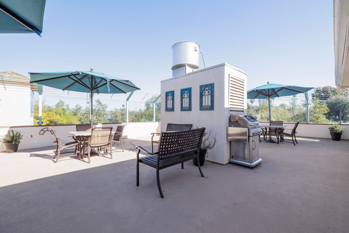 A rooftop patio with seating, umbrellas, and a barbecue grill under a clear blue sky.