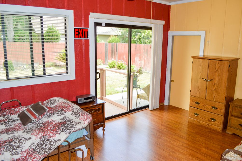 A bright bedroom with a sliding glass door leading to a backyard, featuring a bed, dresser, and wooden flooring.