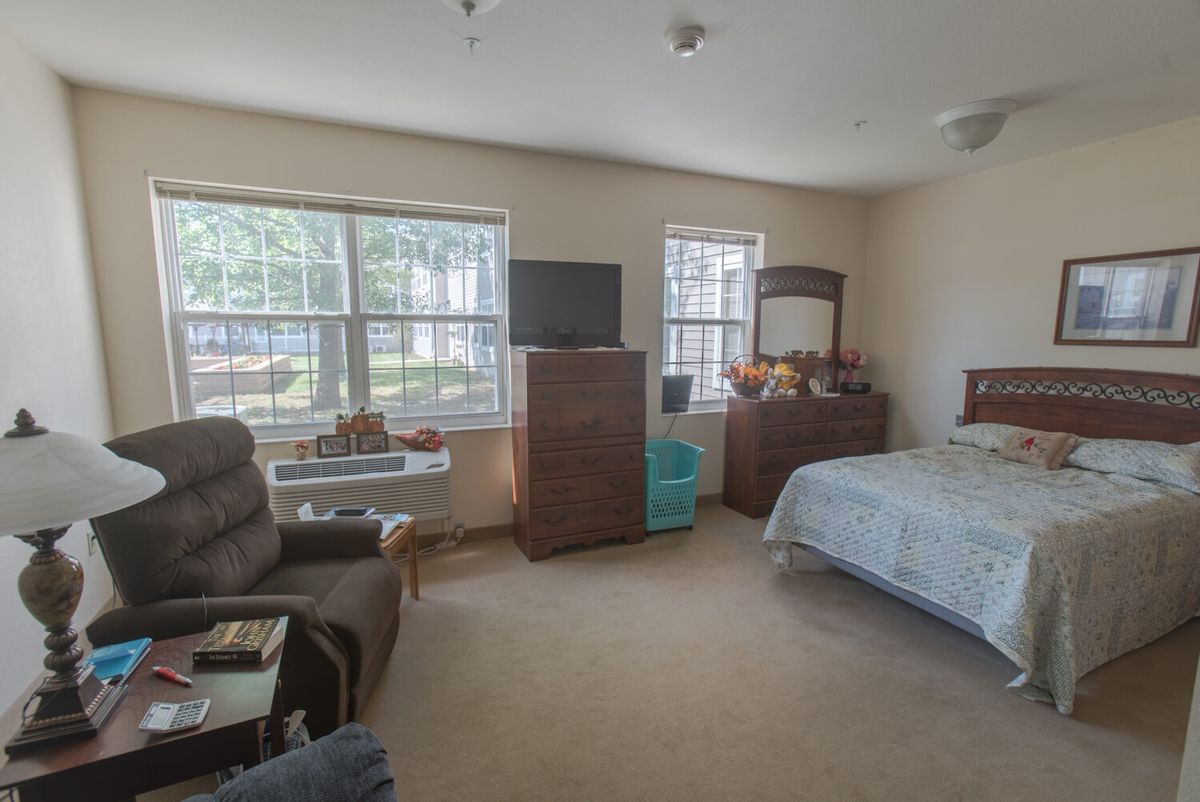 A cozy bedroom with a bed, dresser, TV, and armchair, featuring large windows that let in natural light.
