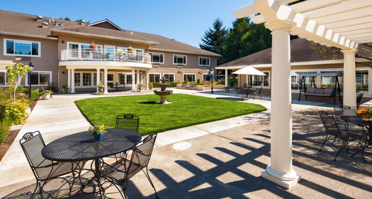 A sunny courtyard with a well-maintained lawn, surrounded by buildings, featuring patio furniture and a central fountain.