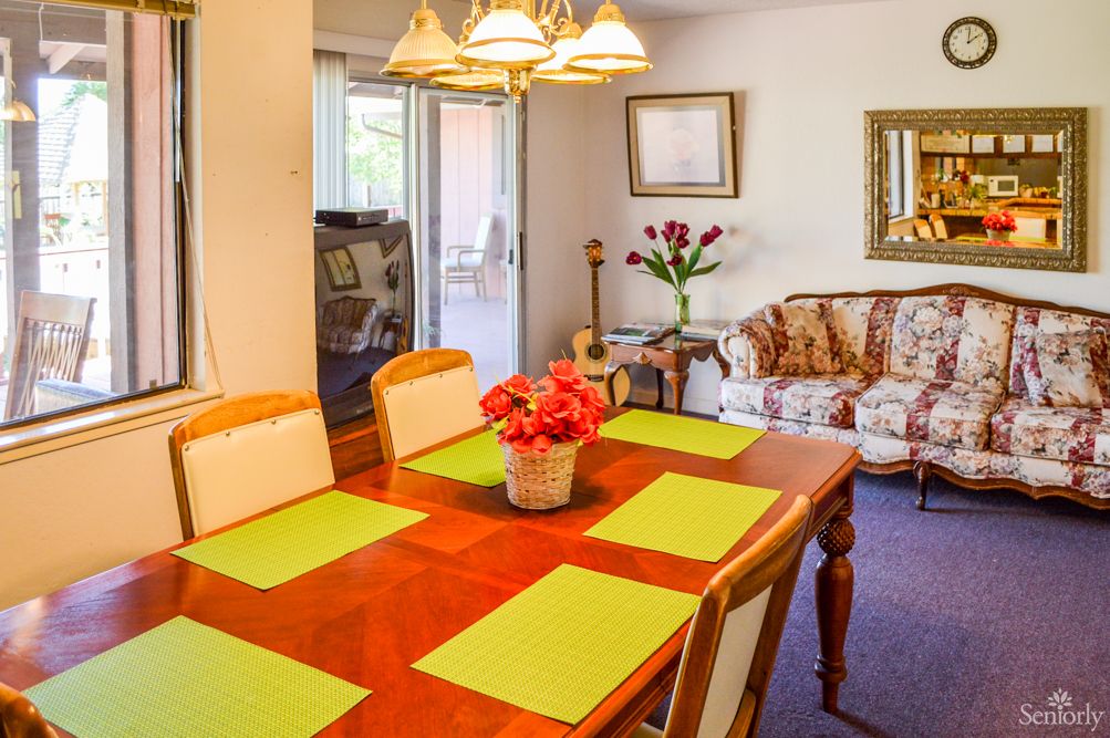A cozy dining and living area featuring a wooden dining table with green placemats, a floral sofa, and decorative elements like flowers and artwork.