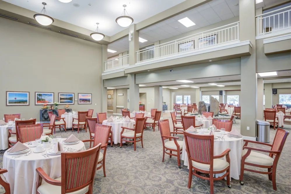 A spacious, elegantly arranged dining room with multiple tables and chairs, featuring an upper-level balcony and stylish lighting fixtures.