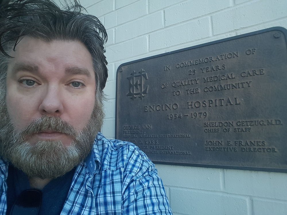 A man with a beard takes a selfie in front of a plaque on a brick wall.
