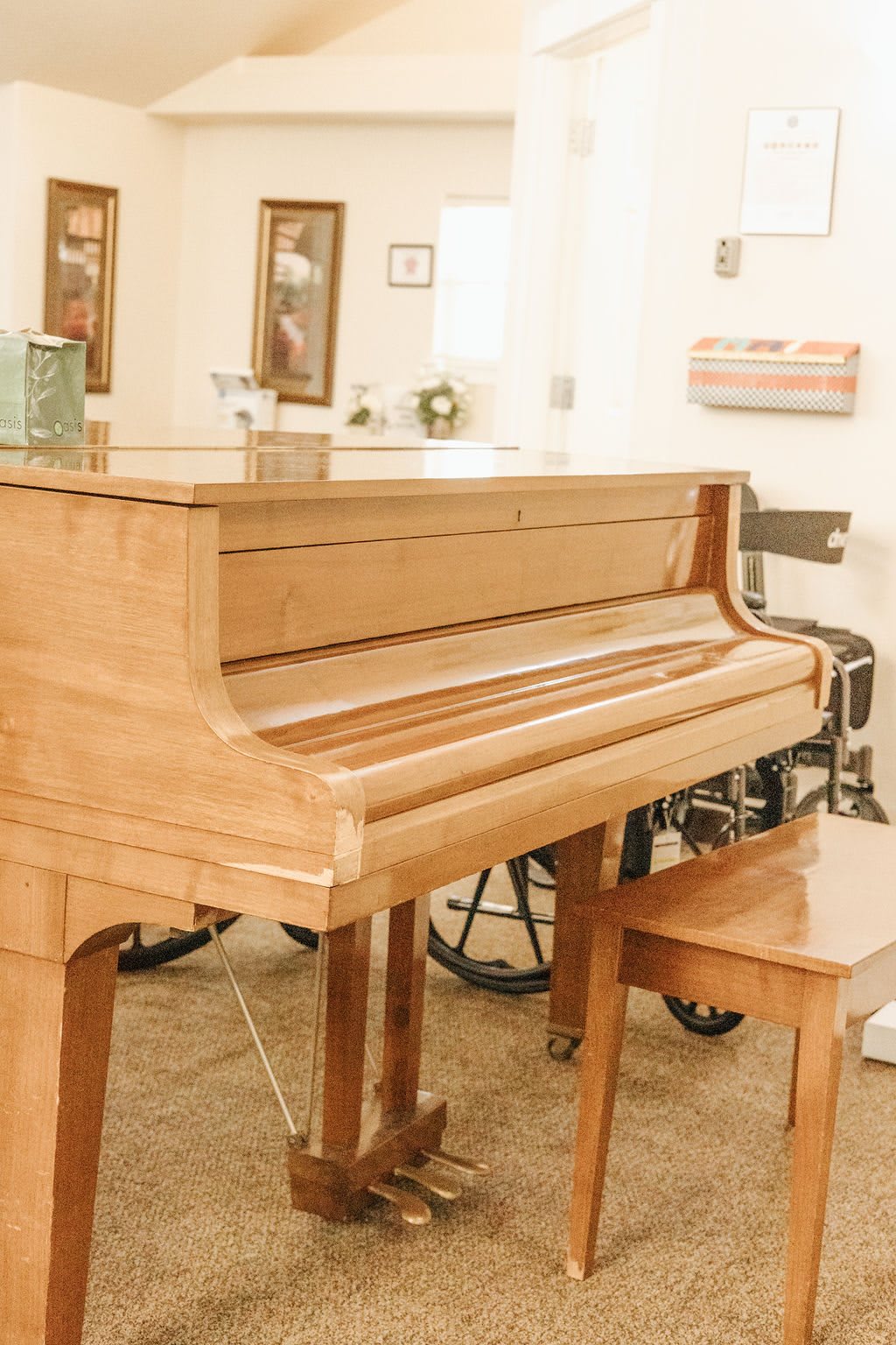 A wooden piano with a matching bench in an indoor setting, accompanied by a wheelchair.