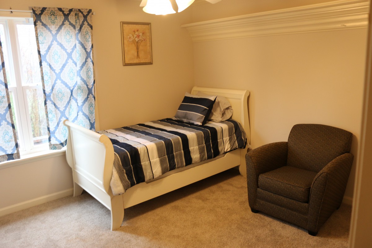 A cozy bedroom featuring a single bed with striped bedding, an armchair, and a window with patterned curtains.