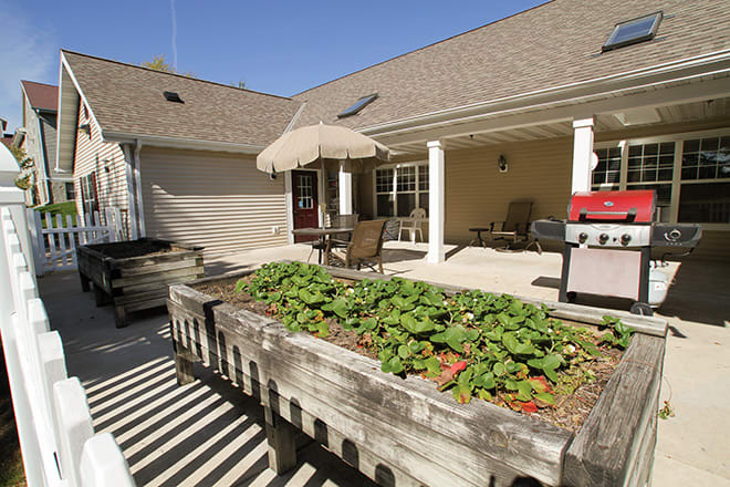 A sunny backyard patio featuring a raised garden bed, outdoor furniture, an umbrella, and a barbecue grill.