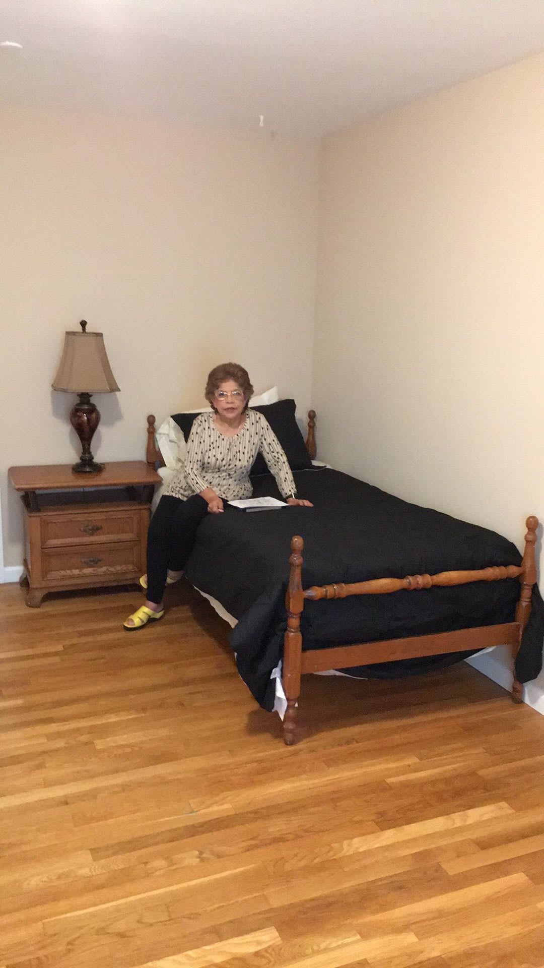 A woman sitting on a bed in a neatly arranged bedroom with wooden flooring.