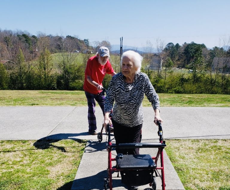 An elderly woman using a walker, accompanied by a man, enjoying a sunny day in the park.