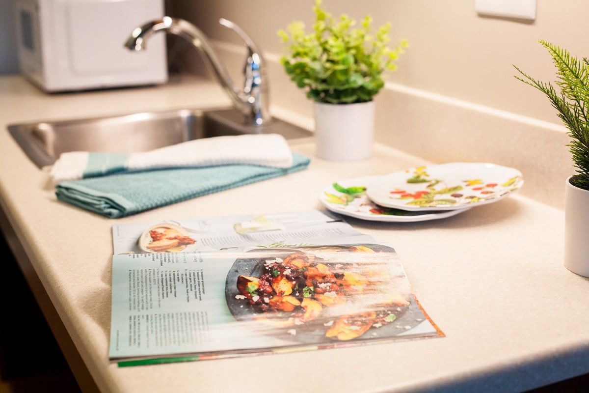 A kitchen countertop with an open recipe book, a small plant, and clean dishes near the sink.
