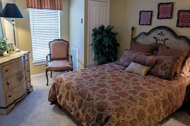 A cozy bedroom with a patterned bedspread, elegant chair, and leafy plant by the window.