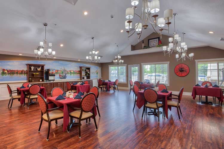 A beautifully decorated dining room with elegant chandeliers, featuring multiple tables and chairs set on a wood floor, with large windows providing natural light.