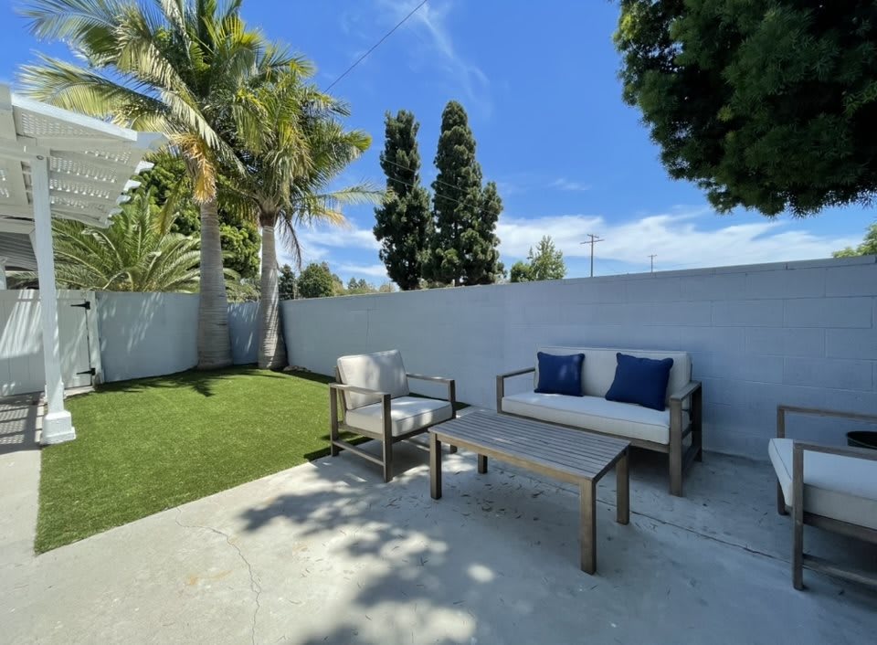 A cozy patio area with seating under a sunny blue sky, surrounded by greenery and palm trees.