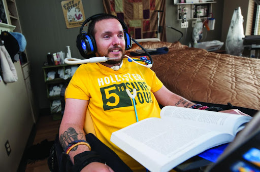 A man in a yellow shirt enjoys gaming using assistive technology while seated in a wheelchair.