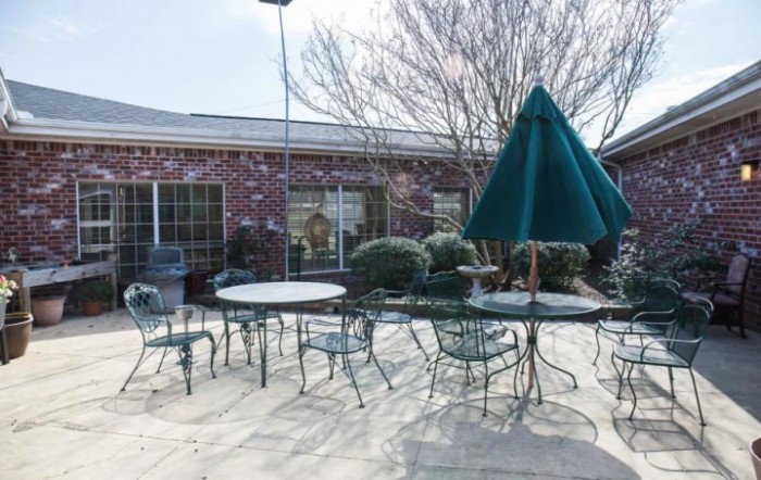 A sunny outdoor patio area with tables, chairs, and a large umbrella beside a brick building.