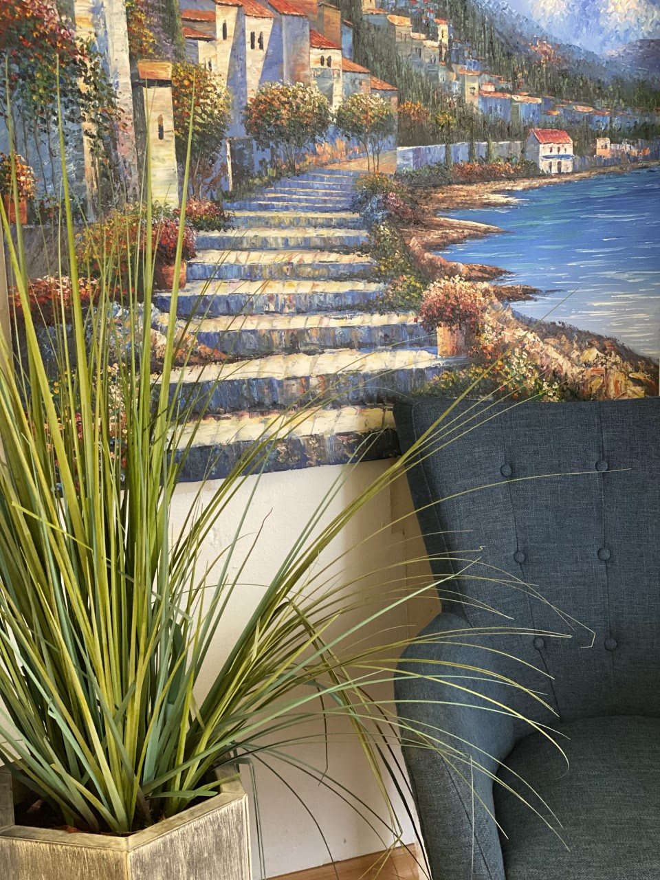 A cozy corner featuring a nature-themed painting with a pathway and coastal scenery, complemented by an indoor plant and a comfortable chair.