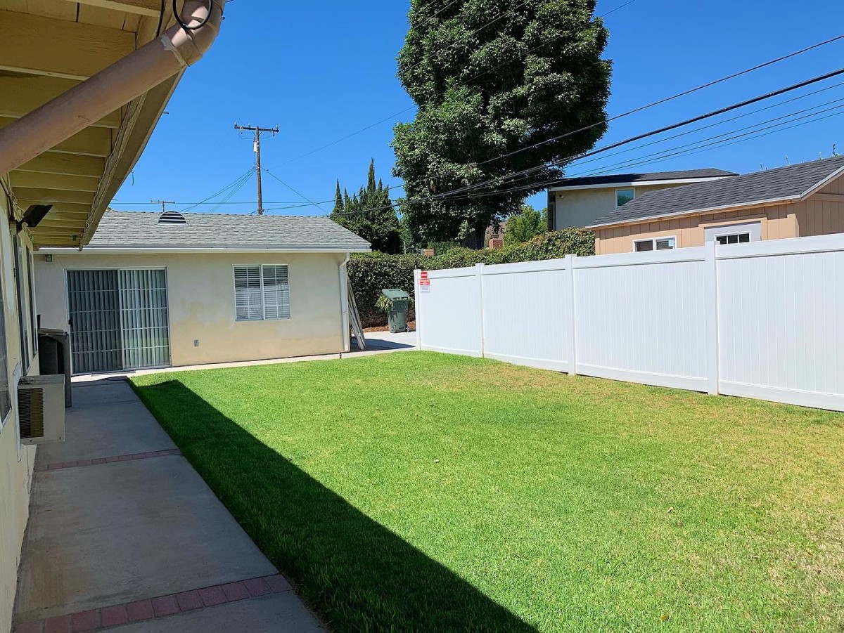 A sunny backyard with a lawn, white fence, and patio area.