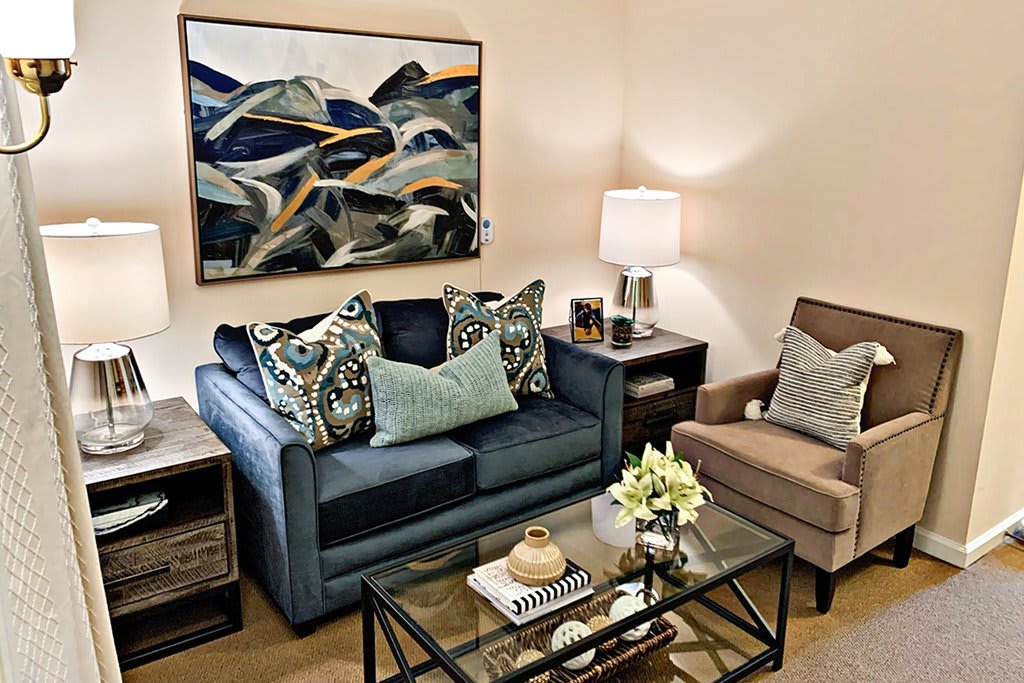 A cozy living room with a modern aesthetic, featuring a blue sofa with decorative pillows, an armchair, and abstract artwork on the wall.