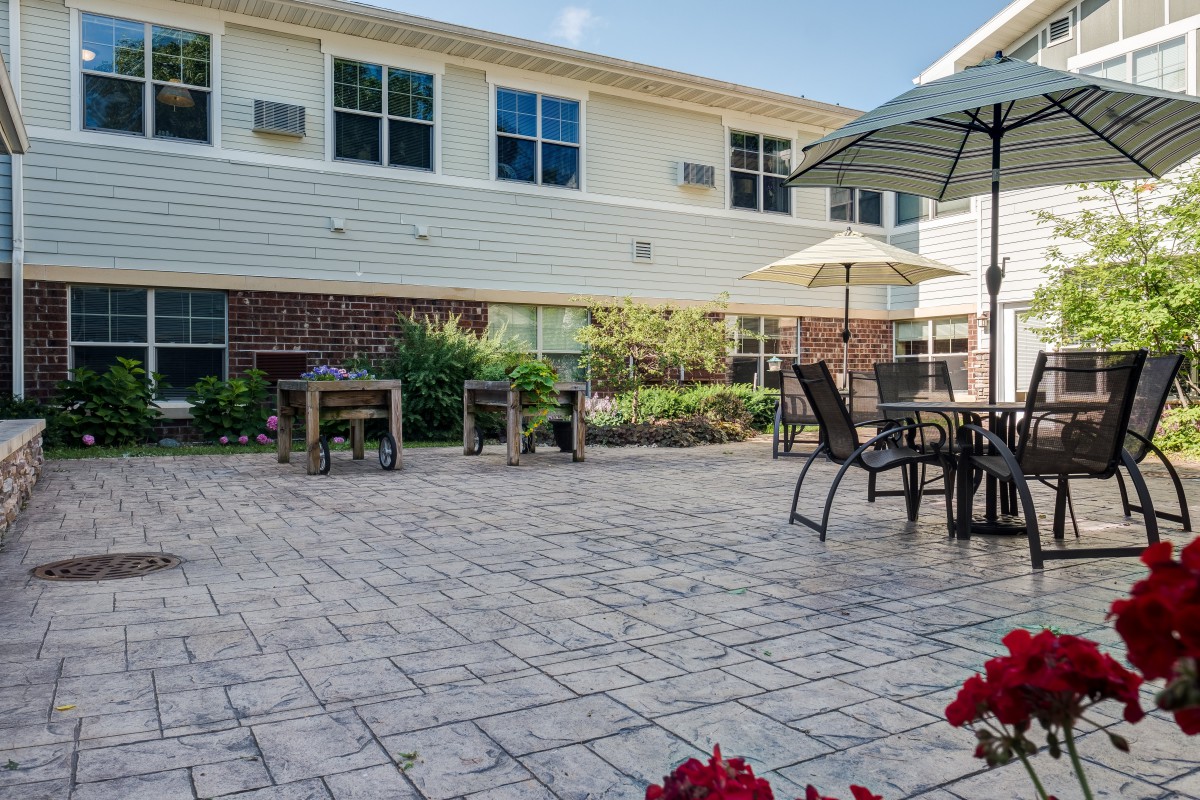 A sunny patio with tables and chairs under umbrellas, surrounded by a garden next to a building.