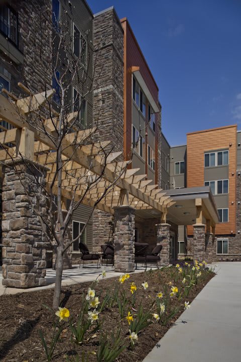 A modern residential building featuring a stone facade and a wooden pergola, surrounded by landscaped greenery.