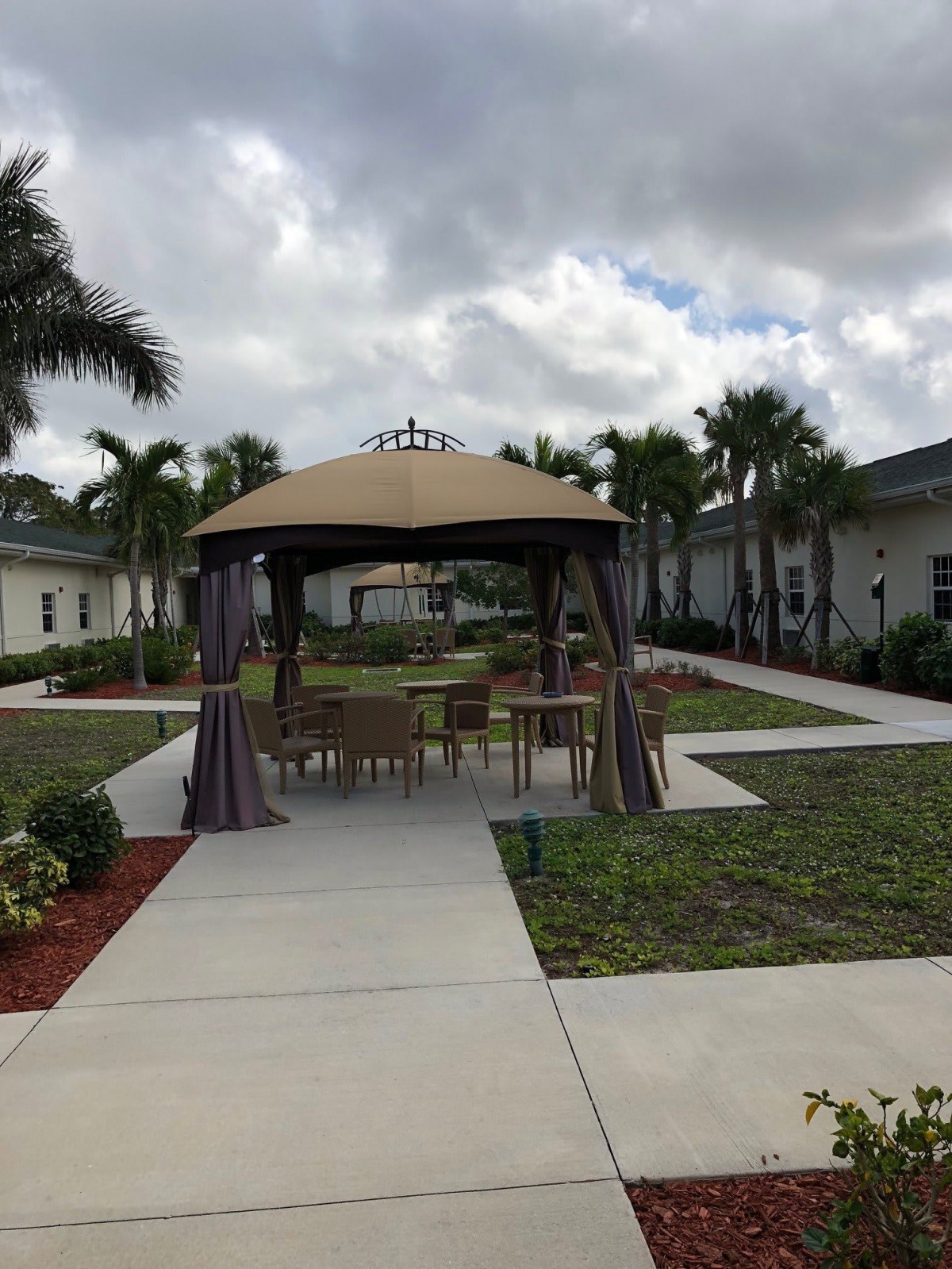 An outdoor area featuring a gazebo surrounded by palm trees and a walkway under a cloudy sky.