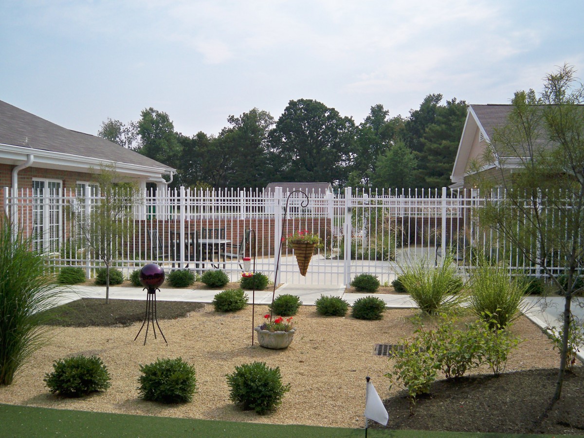 A landscaped garden area with a white fence, various bushes, and plants set against a residential backdrop.