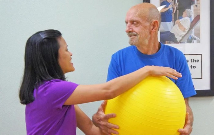A senior man engaging in physical therapy with the assistance of a therapist using a stability ball.
