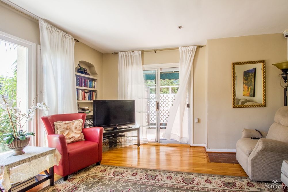 A cozy living room with a red armchair, bookshelves, and a TV sits atop a hardwood floor with a patterned rug, leading to a sliding door covered by sheer curtains.