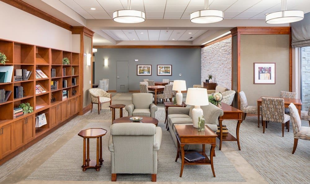 A well-furnished waiting room featuring comfortable chairs, tables, lamps, and a carpeted floor with wood panel accents and bookshelves.