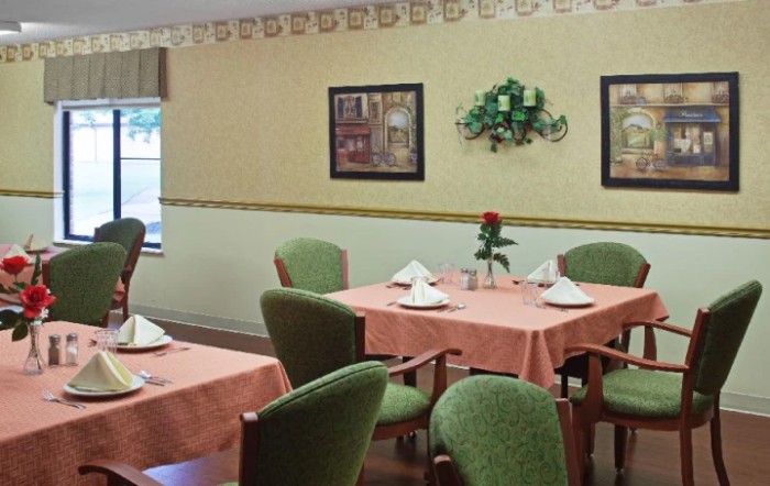 A cozy dining room with neatly set tables, featuring green chairs and floral centerpieces.