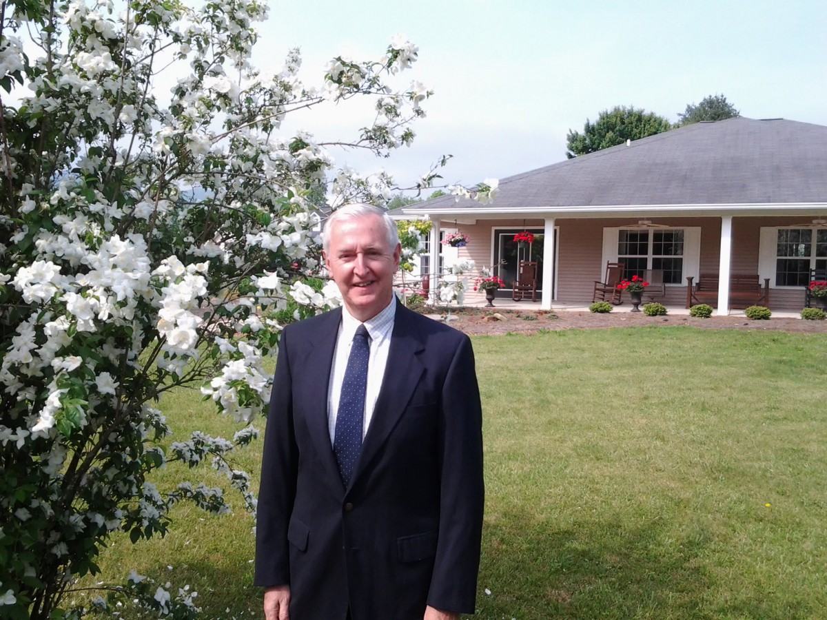 A man in a suit standing outside near flowering plants with a house in the background.