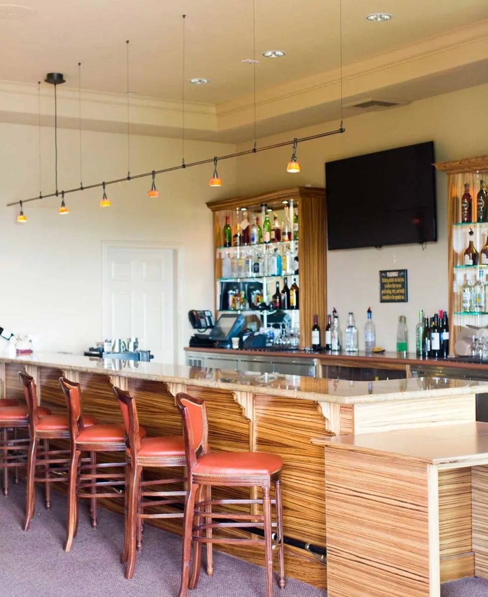 A modern bar with a sleek counter and empty red chairs, featuring a liquor display and a television on the wall.