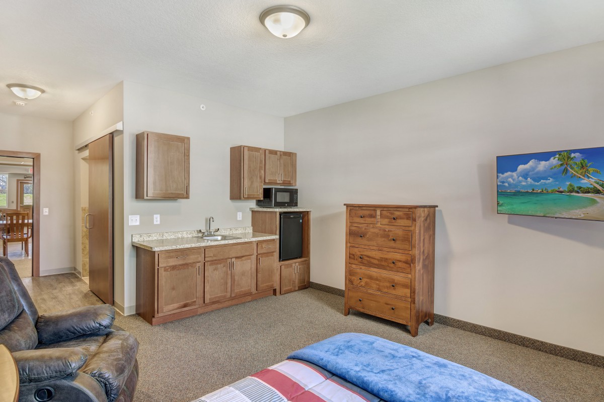 A cozy bedroom with a kitchenette, featuring a microwave and a wooden dresser.