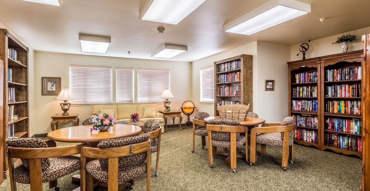 A cozy living room with tables, chairs, bookshelves, and soft lighting.