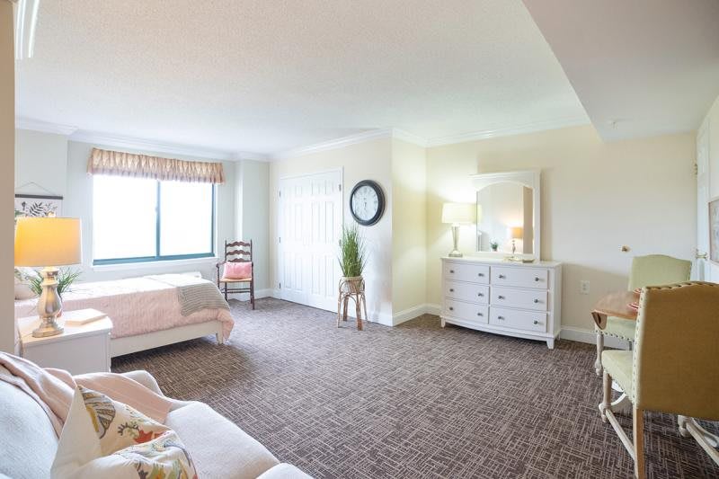 A spacious and bright bedroom with carpeted flooring, a bed, a dresser with a mirror, and a large window.
