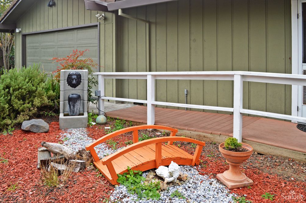 A small decorative garden featuring a mini wooden bridge, gravel pathway, planter, and water fountain next to a wooden deck with railing.