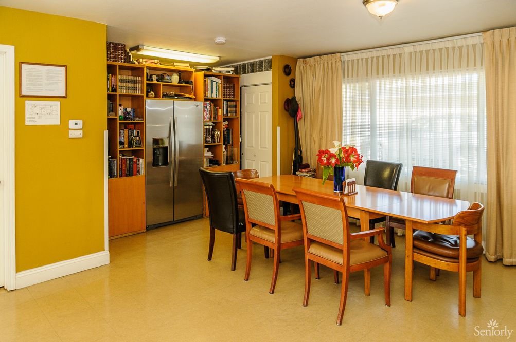 A well-lit dining area with a large table and chairs, adjacent to a kitchen space featuring a stainless steel refrigerator.