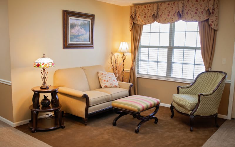 A cozy living room with a beige sofa, armchair, and a small footstool, featuring warm lighting and large windows with curtains.
