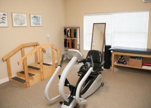A rehabilitation therapy room featuring an elliptical machine, wooden steps, a mirror, and shelves beside a window.